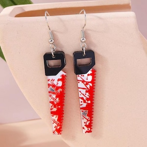 May include: A pair of novelty earrings featuring a saw blade design. The earrings have a black top with a silver hook and a white blade with red splatter details. The saw blade measures approximately 1.5 inches in length.