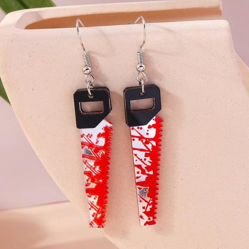 May include: A pair of novelty earrings featuring a saw blade design. The earrings have a black top with a silver hook and a white blade with red splatter details. The saw blade measures approximately 1.5 inches in length.