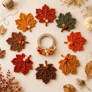 Op de afbeelding: Een verzameling van acht gehaakte esdoornbladeren in diverse herfstkleuren zoals rood, oranje, bruin en groen, elk versierd met kleine gehaakte besjes. Een ronde gehaakte krans met een trosje besjes bevindt zich in het midden.