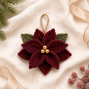 May include: A crocheted poinsettia ornament featuring deep burgundy petals and dark green leaves. The center of the flower is decorated with three small, shiny gold beads. A natural jute twine loop is attached to the top for hanging purposes.