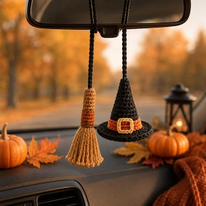 Puede incluir: Un accesorio de coche tejido a ganchillo que cuelga del espejo retrovisor, con un pequeño sombrero de bruja negro con banda y hebilla naranja, junto a una escoba de color beige y naranja, ambos suspendidos por cordones trenzados negros sobre un fondo otoñal.