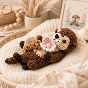 Puede incluir: Un peluche de nutria tejido a ganchillo en color marrón con vientre crema, patas marrón oscuro y ojos de seguridad negros. La nutria sostiene una pequeña concha rosa tejida con una perla blanca, con una nutria bebé unida a su pecho.
