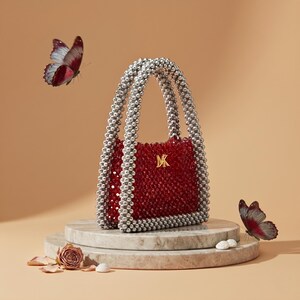 May include: A red beaded handbag with silver beaded handles and a gold "MK" emblem. The bag is on a marble pedestal, with butterflies and a rose. The bag's dimensions are not visible.