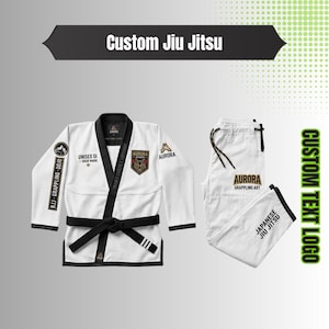 Könnte beinhalten: Ein weißes Jiu-Jitsu-Gi-Set bestehend aus einer Jacke mit schwarzem Besatz, einem schwarzen Gürtel und einer passenden Hose. Die Jacke zeigt den Text "Custom Jiu Jitsu", "Unisex Gi", "Krav Maga", "BJJ Grappling Dojo" und "Aurora Academy" mit einem Wolfslogo. Die Hose ist mit "Aurora Grappling Art" und "Japanese Jiu Jitsu" beschriftet.