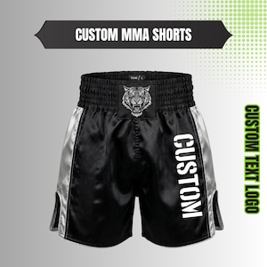 May include: Black MMA shorts with silver side panels and a wide elastic waistband. A tiger head graphic is centered on the front waistband. The word CUSTOM is printed vertically in white on the right leg. The text CUSTOM MMA SHORTS and CUSTOM TEXT LOGO appear.