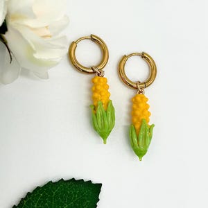 May include: A pair of gold-toned hoop earrings featuring dangling charms shaped like ears of corn. Each charm has a bright yellow textured kernel section and light green husks at the base, suspended from a simple circular metal hoop closure.