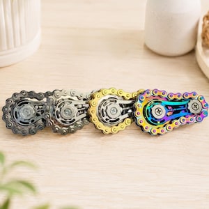 May include: A set of four fidget toys constructed from metal bicycle chain links and gears. The set includes one dark grey, one silver, one gold, and one iridescent rainbow-coloured toy, each featuring two central screws and a mechanical design.