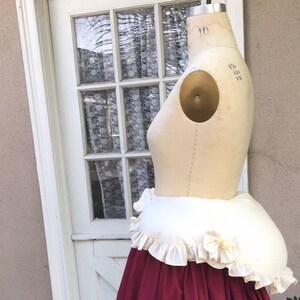 Puede incluir: Un enagua blanca y crema con ribete de volantes, puesta en un maniqu&iacute;. La enagua es de forma redonda y grande y est&aacute; dise&ntilde;ada para dar un aspecto lleno y redondeado a la parte posterior de una falda.