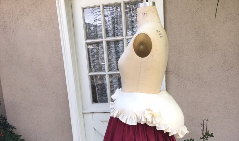 Puede incluir: Un enagua blanca y crema con ribete de volantes, puesta en un maniqu&iacute;. La enagua es de forma redonda y grande y est&aacute; dise&ntilde;ada para dar un aspecto lleno y redondeado a la parte posterior de una falda.