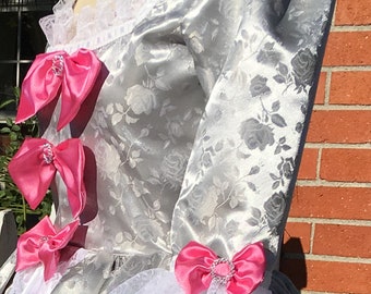 May include: A silver satin dress with a white lace trim and pink satin bows. The dress has a floral pattern.