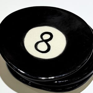 Può includere: Una pila di piatti rotondi in ceramica nera con finitura lucida. Ogni piatto presenta un inserto circolare bianco al centro contenente un numero otto nero dipinto a mano, che imita l'aspetto di una classica palla da biliardo.