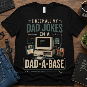 May include: Black t-shirt with the text "I KEEP ALL MY DAD JOKES IN A DAD-A-BASE". The design includes a computer, floppy disks, and a database icon. The shirt is on a wooden surface with other items.