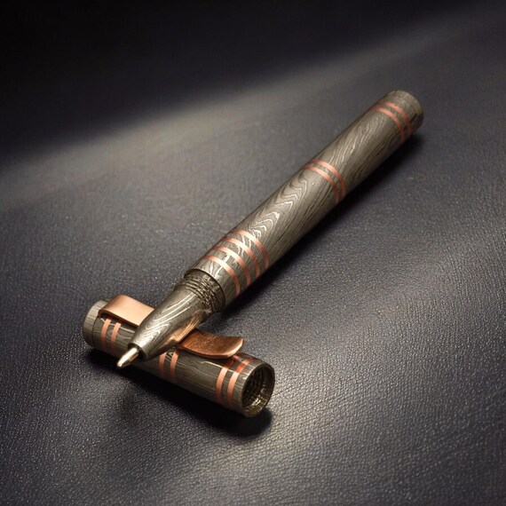 A hand-forged Damascus steel bullet pen with copper inlay — this is actual blacksmithing craft meeting everyday carry, the kind of object that stops people mid-sentence to ask 'wait, what IS that?'