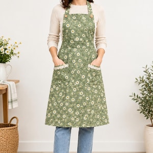 Puede incluir: Un delantal de color verde salvia con un patrón repetitivo de margaritas blancas, dos bolsillos delanteros con ribete de encaje blanco y dos botones de madera en la parte superior donde se sujetan las correas. Cubre el torso hasta los muslos.