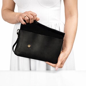 Peut inclure: Une pochette en cuir noir avec une silhouette rectangulaire, un rabat repliable doté d'un bouton-pression doré et une dragonne latérale. L'avant du sac présente un logo subtilement gaufré dans le coin inférieur gauche.