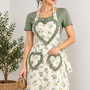 Peut inclure: Un tablier couleur crème orné d'un motif floral composé de petites fleurs bleues et jaunes, agrémenté de trois poches en forme de cœur bordées de volants vert sauge. Le tablier présente une bordure en dentelle blanche et des liens vert sauge.