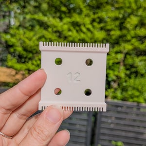 Peut inclure: Lame de tondeuse à cheveux beige clair, portant le numéro 12. La lame est rectangulaire, avec de petites dents sur les bords supérieur et inférieur. Elle comporte quatre trous circulaires. L'arrière-plan est une vue floue de feuillage vert.