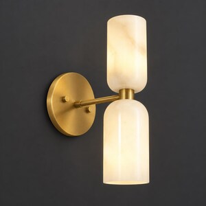 May include: A wall sconce featuring a circular brushed brass backplate and a horizontal arm extending to two vertically stacked, cylindrical, translucent white stone shades that emit a soft, warm glow from within the light fixture.