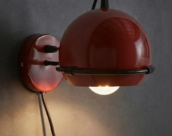 Pode incluir: Um candeeiro de parede com um abajur vermelho brilhante em forma de cúpula, sustentado por uma estrutura de metal preto. O candeeiro inclui uma placa de parede circular vermelha, um cabo de alimentação preto e uma lâmpada acesa.