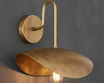 Pode incluir: Um candeeiro de parede com uma placa circular de latão e um braço curvo que suporta um abajur de latão largo e raso em forma de pires. Uma lâmpada de vidro transparente pende para baixo a partir do centro do abajur metálico.