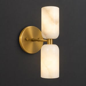 May include: A wall-mounted light fixture featuring a circular brass backplate and a horizontal arm that supports two vertically stacked, cylindrical, translucent white stone shades that emit a soft, warm glow from within the stone structure.