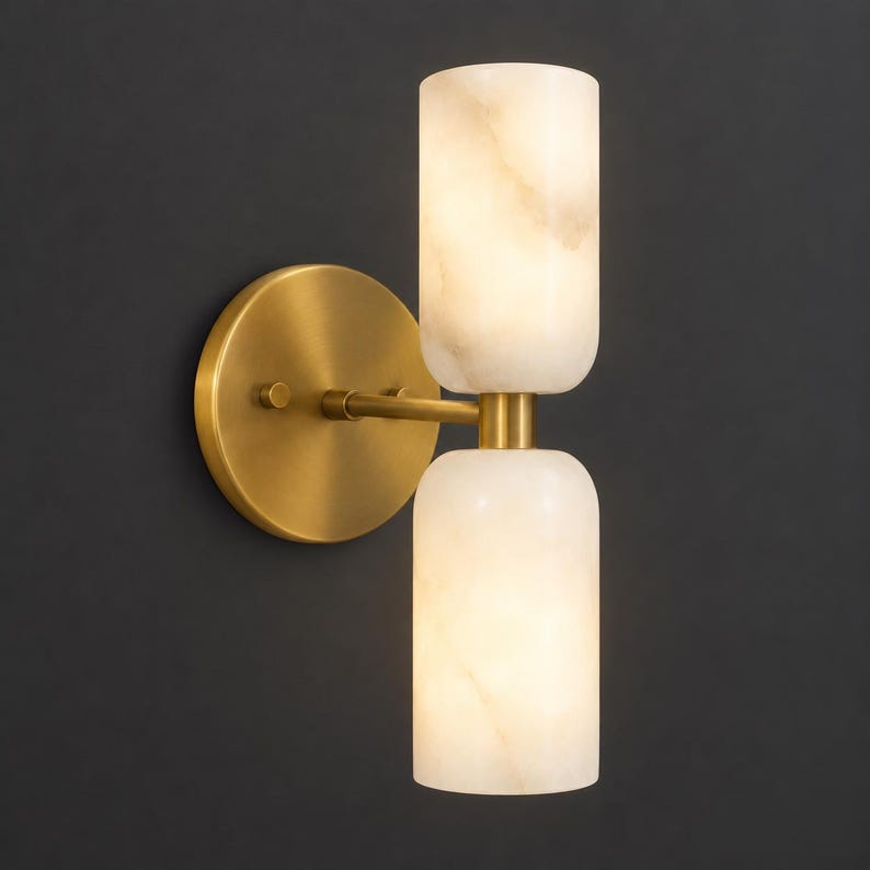 May include: A wall-mounted light fixture featuring a circular brass backplate and a horizontal arm that supports two vertically stacked, cylindrical, translucent white stone shades that emit a soft, warm glow from within the stone structure.
