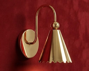 Pode incluir: Um candeeiro de parede em tom dourado com uma placa de montagem circular, um braço curvo e um abajur cónico com uma extremidade inferior recortada. O candeeiro tem um acabamento metálico polido e refletor com uma peça esférica de ligação.