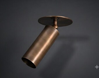 Pode incluir: Um foco cilíndrico com acabamento em bronze escovado. A luminária possui uma placa de montagem circular ligada ao tubo principal por uma junta giratória, permitindo o posicionamento ajustável da fonte de luz numa direção descendente.