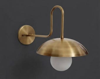 Pode incluir: Um candeeiro de parede com acabamento em latão escovado. O design inclui uma placa de parede circular, um braço curvo que se estende para fora e uma cúpula de metal larga posicionada sobre uma lâmpada esférica de vidro fosco branco.