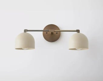 Pode incluir: Uma luminária de parede com uma placa de fixação circular em bronze e uma barra horizontal que suporta duas cúpulas em formato de domo na cor creme fosco. O design inclui lâmpadas visíveis dentro de cada cúpula para iluminação direta.