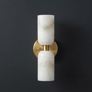 May include: A wall sconce featuring two cylindrical white stone shades with subtle beige veining, connected by a central brass-toned metal post and mounted on a circular brass-toned backplate, creating a vertical, symmetrical lighting fixture.