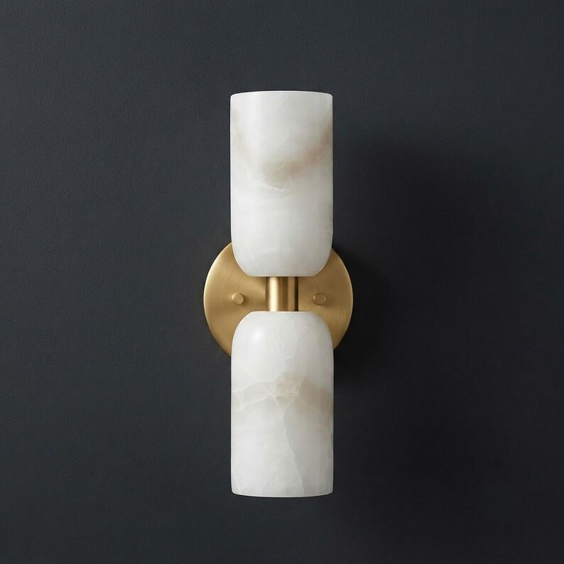 May include: A wall sconce featuring two cylindrical white stone shades with subtle beige veining, connected by a central brass-toned metal post and mounted on a circular brass-toned backplate, creating a vertical, symmetrical lighting fixture.