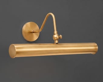 Pode incluir: Uma luminária de parede para quadros com acabamento em latão escovado. A peça apresenta uma base circular, um braço curvo e um corpo cilíndrico horizontal para a luz, com uma articulação ajustável e um pequeno botão lateral de ajuste.