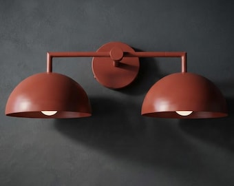 Pode incluir: Uma luminária de parede com acabamento em terracota fosco. O design inclui uma placa de montagem circular central conectada a dois braços horizontais, cada um sustentando uma cúpula voltada para baixo com uma lâmpada individual.