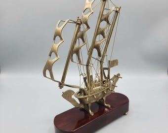 Pode incluir: Um modelo de navio de metal na cor latão com velas recortadas estilizadas e linhas de cordame finas. A embarcação está montada sobre uma base de madeira oval lisa de cor castanho-escuro com pés pequenos, exibindo uma estrutura de casco detalhada.