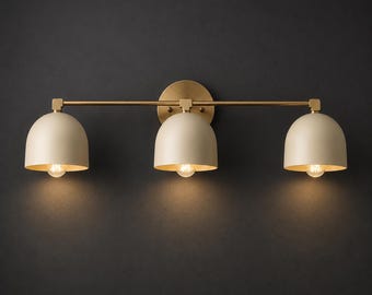 Pode incluir: Um candeeiro de parede de três luzes com uma barra horizontal de latão e uma placa de parede circular de latão. Três cúpulas de cor creme mate estão fixadas à barra, cada uma contendo uma lâmpada que emite uma luz quente e direta.