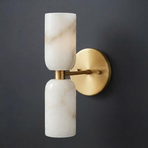 May include: A wall sconce featuring two cylindrical white stone shades with subtle grey veining, stacked vertically and connected by a central brass arm to a circular brass wall plate. The fixture has a polished metallic finish and stone texture.