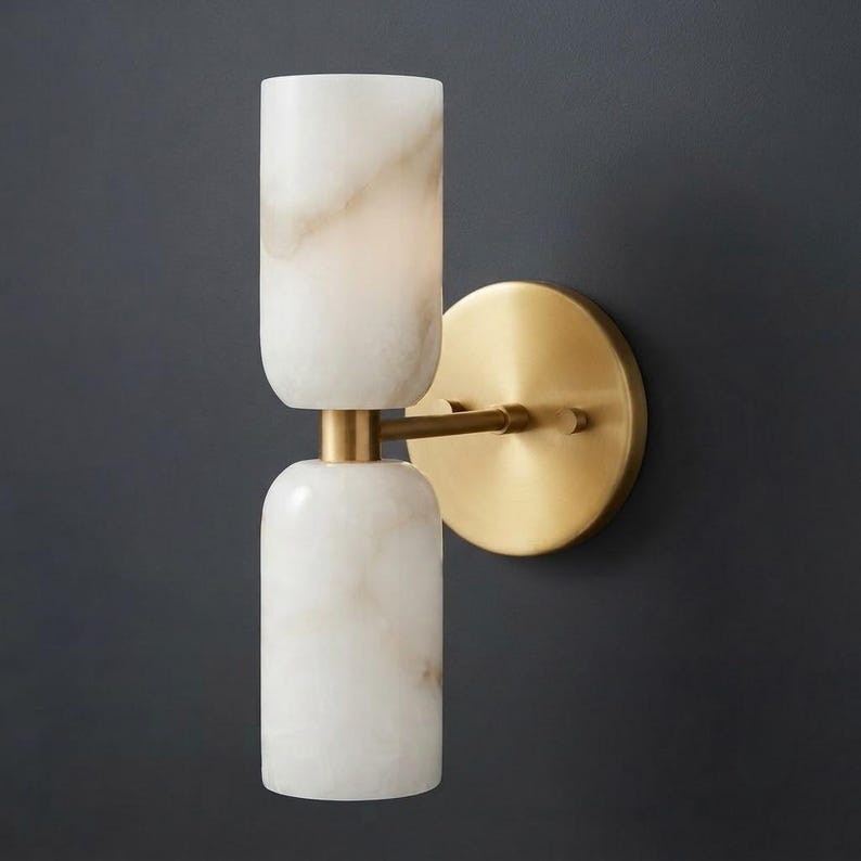 May include: A wall sconce featuring two cylindrical white stone shades with subtle grey veining, stacked vertically and connected by a central brass arm to a circular brass wall plate. The fixture has a polished metallic finish and stone texture.