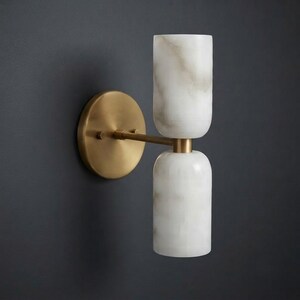 May include: A wall sconce featuring a circular brass backplate and a horizontal arm extending to two vertically stacked, cylindrical white marble shades with grey veining. The fixture has a metallic finish and a smooth, polished appearance.