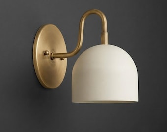 Pode incluir: Um candeeiro de parede com uma placa circular de latão e um braço curvo de latão que suporta um abajur em forma de cúpula de cor branco-sujo. Os componentes metálicos têm um acabamento escovado e o abajur direciona a luz para baixo.