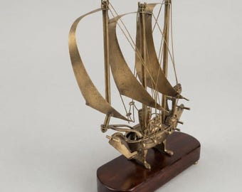 Pode incluir: Um modelo de veleiro em latão com dois mastros, velas de metal curvas e cordame de arame fino. O navio está montado sobre uma base de madeira oval de cor castanho escuro com pequenos pés, exibindo um acabamento metálico detalhado.