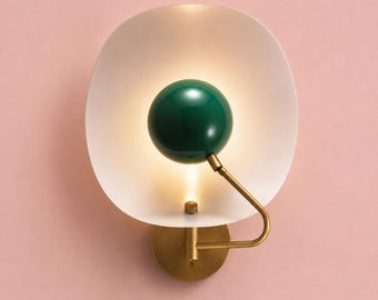 Pode incluir: Um candeeiro de parede com um grande abajur em forma de taça de cor branco-sujo e uma luminária central esférica de cor verde-escura brilhante. O candeeiro é suportado por um braço metálico cor de latão e uma base circular, emitindo uma luz quente.
