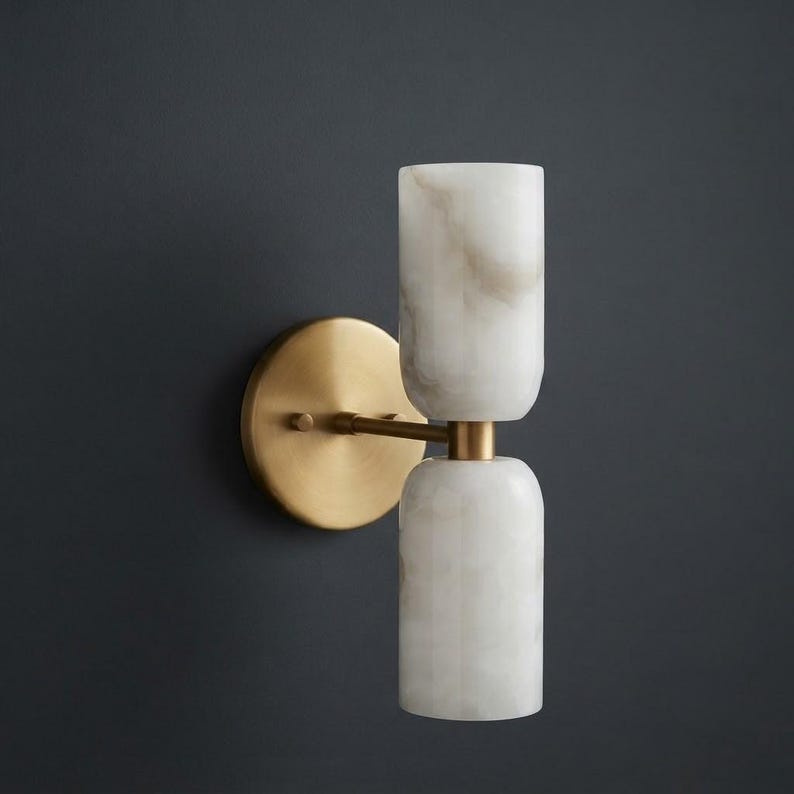 May include: A wall sconce featuring a circular brass backplate and a horizontal arm supporting two vertically stacked, cylindrical white stone shades with subtle grey veining. The fixture has a metallic finish and a clean, symmetrical design.