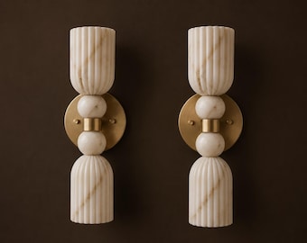 May include: A pair of wall sconces featuring vertical fluted off-white marble shades and central marble spheres connected by a brushed brass cylindrical spacer. Each fixture is mounted on a circular brass backplate with two small brass screws.