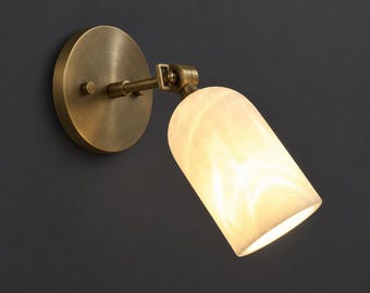 Pode incluir: Um candeeiro de parede com uma placa de montagem circular em latão antigo e um braço ajustável ligado a um abajur cilíndrico de alabastro branco translúcido que emite um brilho quente e suave para iluminação direcional em interiores.