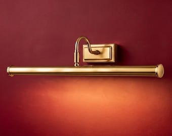 Pode incluir: Uma luminária de quadro em latão polido com base retangular de parede, braço curvo e uma cabeça de lâmpada cilíndrica horizontal longa que projeta um brilho quente voltado para baixo na superfície da parede atrás da luminária.