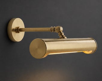 Pode incluir: Uma luminária de parede para quadros com acabamento em latão escovado. O dispositivo possui uma placa de parede circular, uma cabeça de lâmpada cilíndrica horizontal e um braço articulado ajustável com um mecanismo de dobradiça visível que emite uma luz quente.