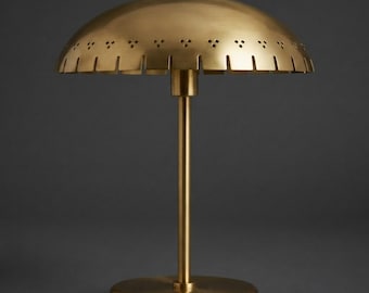 Pode incluir: Um candeeiro de mesa em latão com um abajur largo em forma de cúpula, uma borda recortada e uma série de pequenos recortes triangulares ao longo do perímetro. O candeeiro tem uma haste central fina e uma base circular em latão mate.