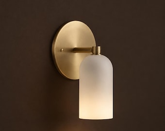 Pode incluir: Um candeeiro de parede com uma placa circular de latão escovado e um braço cilíndrico que sustenta um abajur vertical de vidro branco fosco. A luminária emite um brilho suave e quente através do vidro, criando um visual equilibrado.