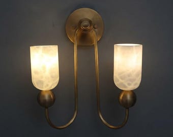 May include: A double wall sconce featuring a circular brass backplate and two curved arms extending downward. Each arm supports a spherical brass base topped with a cylindrical, translucent white glass shade that emits a soft, warm glow.